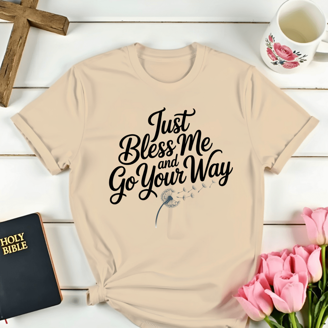 Beige t-shirt with 'Just Bless Me and Go Your Way' text on a wooden surface with flowers and a mug.