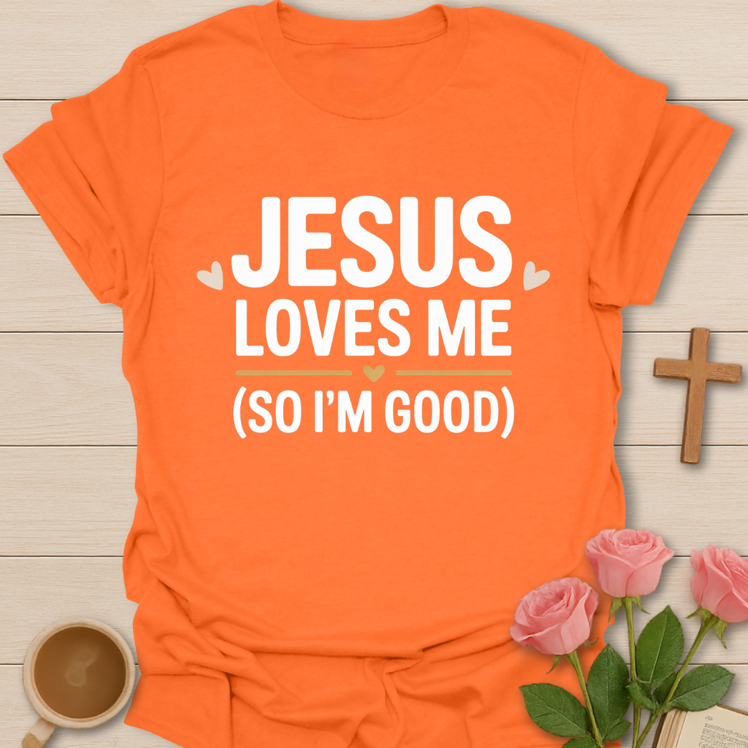 Orange t-shirt with 'Jesus loves me (so I'm good)' text on a wooden surface with flowers and a cross.