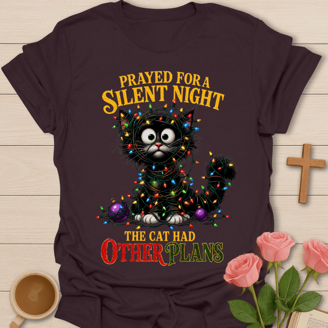 A dark brown graphic t-shirt laid flat on a light wood background, decorated with a Christmas-themed design featuring a black cat wrapped in colorful lights and the text 'PRAYED FOR A SILENT NIGHT THE CAT HAD OTHER PLANS'. The t-shirt is styled with pink roses, a wooden cross, and a cup of coffee.