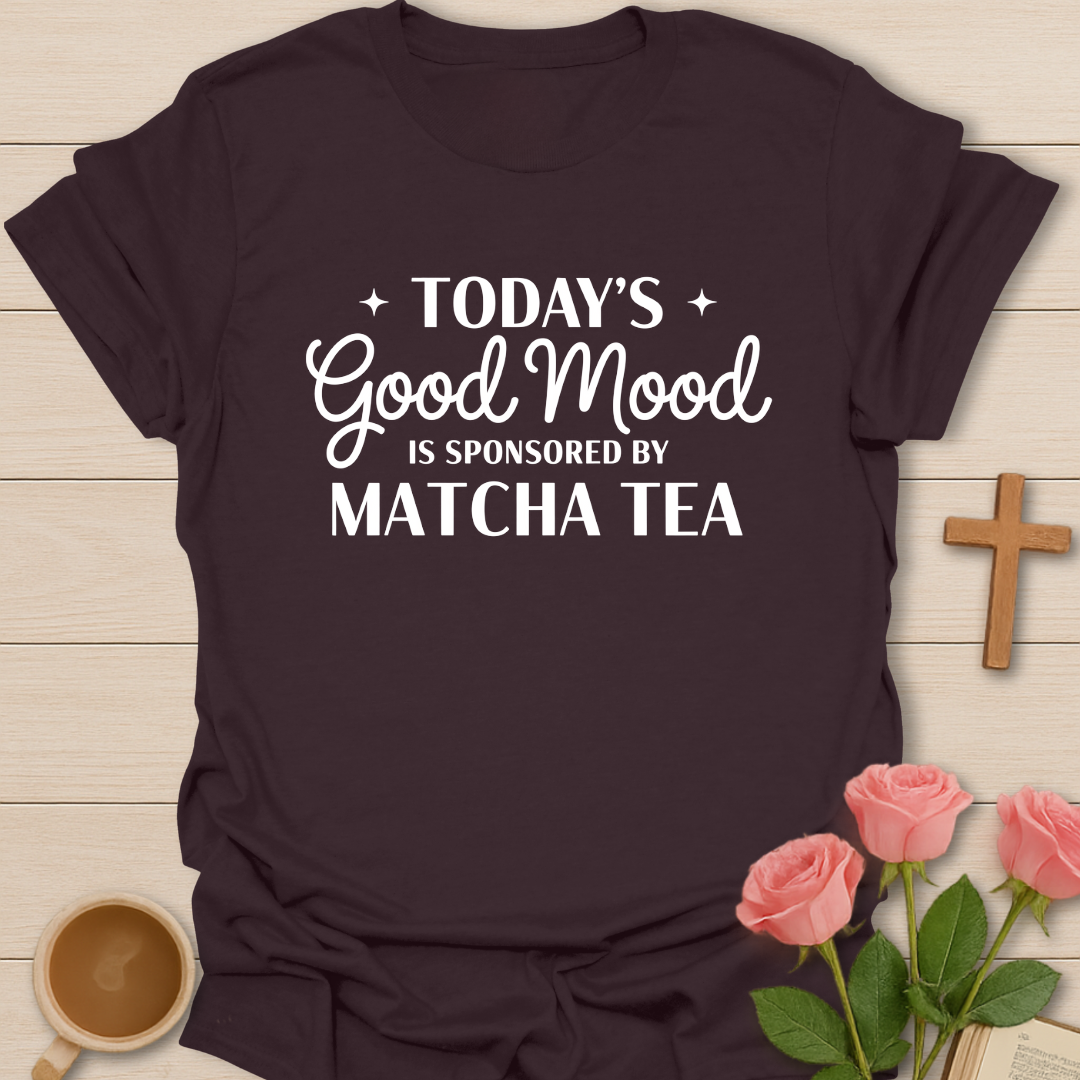 Brown t-shirt with 'Today's Good Mood is Sponsored by Matcha Tea' text on a wooden surface with flowers and a cup.