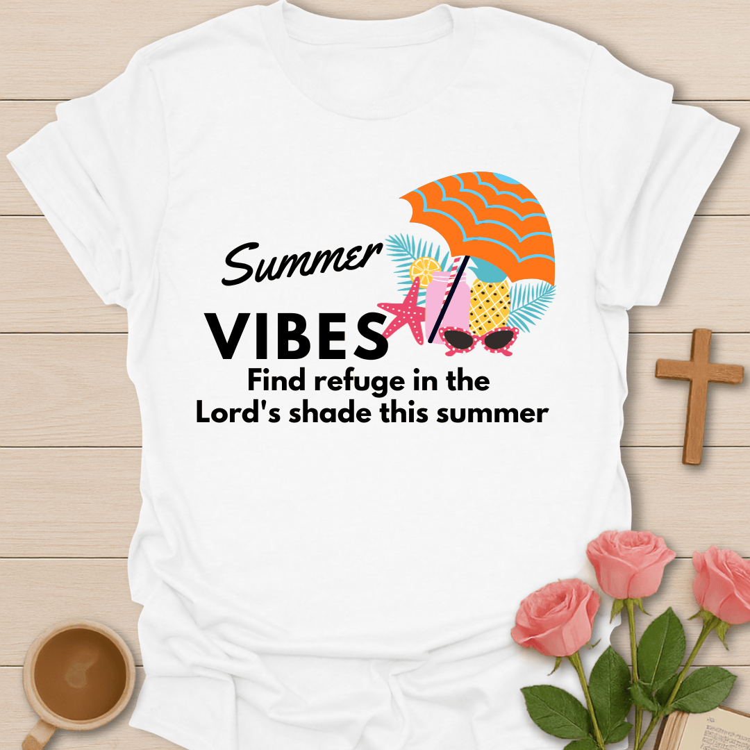 A white short-sleeve T-shirt laid flat on a light wood surface, printed with the phrase 'Summer VIBES Find refuge in the Lord's shade this summer' and a colorful beach-themed graphic including an orange striped umbrella, starfish, palm leaves, and sunglasses. Pink roses, a wooden cross, and a cup are placed around the shirt as styling props.