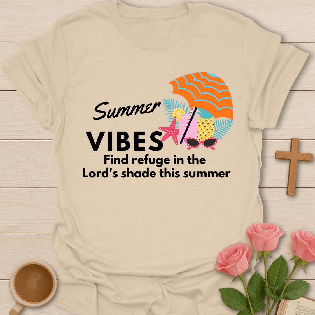 A beige short-sleeve T-shirt laid flat on a light wood surface, printed with the words 'Summer VIBES Find refuge in the Lord's shade this summer' alongside a colorful beach-themed graphic including an orange striped umbrella, pink starfish, sunglasses, and palm leaves. Decorative props around it include pink roses, a wooden cross, and a cup of coffee.