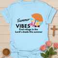 A light blue short-sleeve t-shirt laid flat on a light wood background, featuring a 'Summer Vibes' graphic with an orange beach umbrella, starfish, pineapple, sunglasses, and the text 'Find refuge in the Lord's shade this summer.' Decorative pink roses, a wooden cross, and a coffee cup surround the shirt.