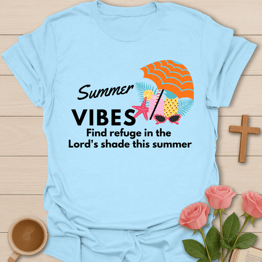 A light blue short-sleeve t-shirt laid flat on a light wood background, featuring a 'Summer Vibes' graphic with an orange beach umbrella, starfish, pineapple, sunglasses, and the text 'Find refuge in the Lord's shade this summer.' Decorative pink roses, a wooden cross, and a coffee cup surround the shirt.