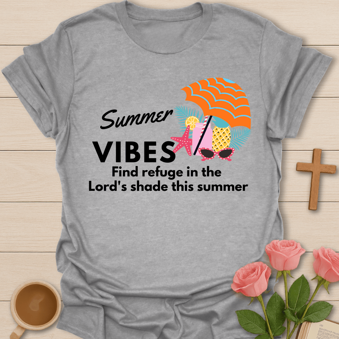 A heather gray short-sleeve t-shirt laid flat on a light wood surface, featuring a colorful summer graphic with the text 'Summer VIBES Find refuge in the Lord's shade this summer' and an umbrella, starfish, and sunglasses design. Pink roses, a coffee cup, and a wooden cross are styled around the shirt.