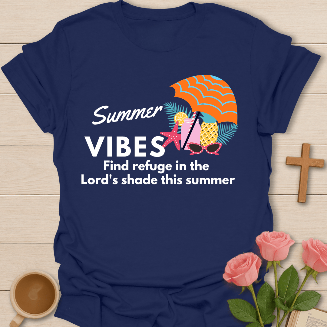 A navy blue graphic t-shirt laid flat on a light wood background, surrounded by pink roses, a wooden cross, and a cup of coffee. The t-shirt features a colorful graphic of pineapples under a striped orange umbrella with the text 'Summer VIBES Find refuge in the Lord's shade this summer'.