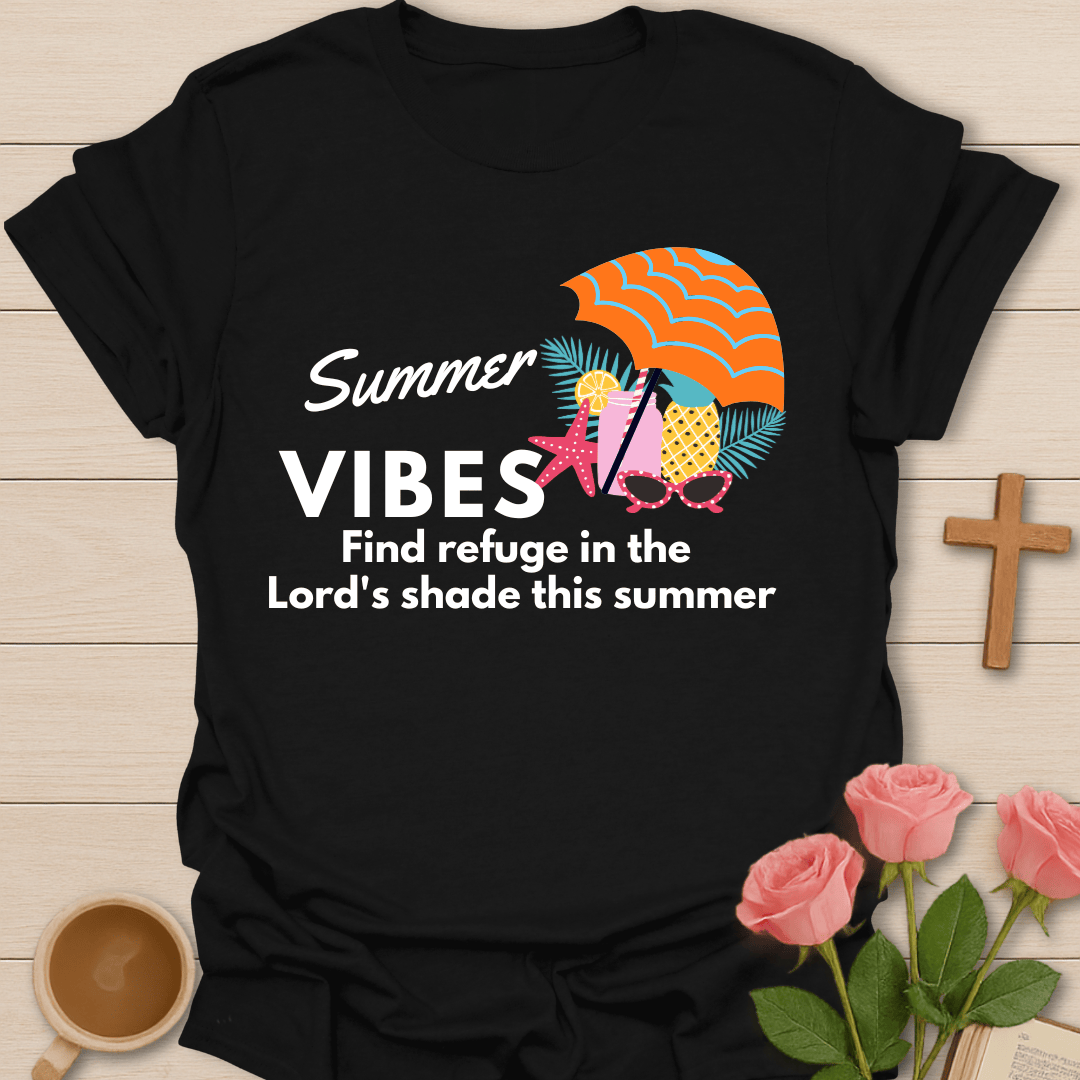 A black short-sleeve graphic T-shirt laid flat on a light wood surface, with a colorful summer-themed print reading “Summer VIBES Find refuge in the Lord’s shade this summer.” The design includes an orange striped umbrella, pink starfish, yellow pineapple, and teal palm leaves.