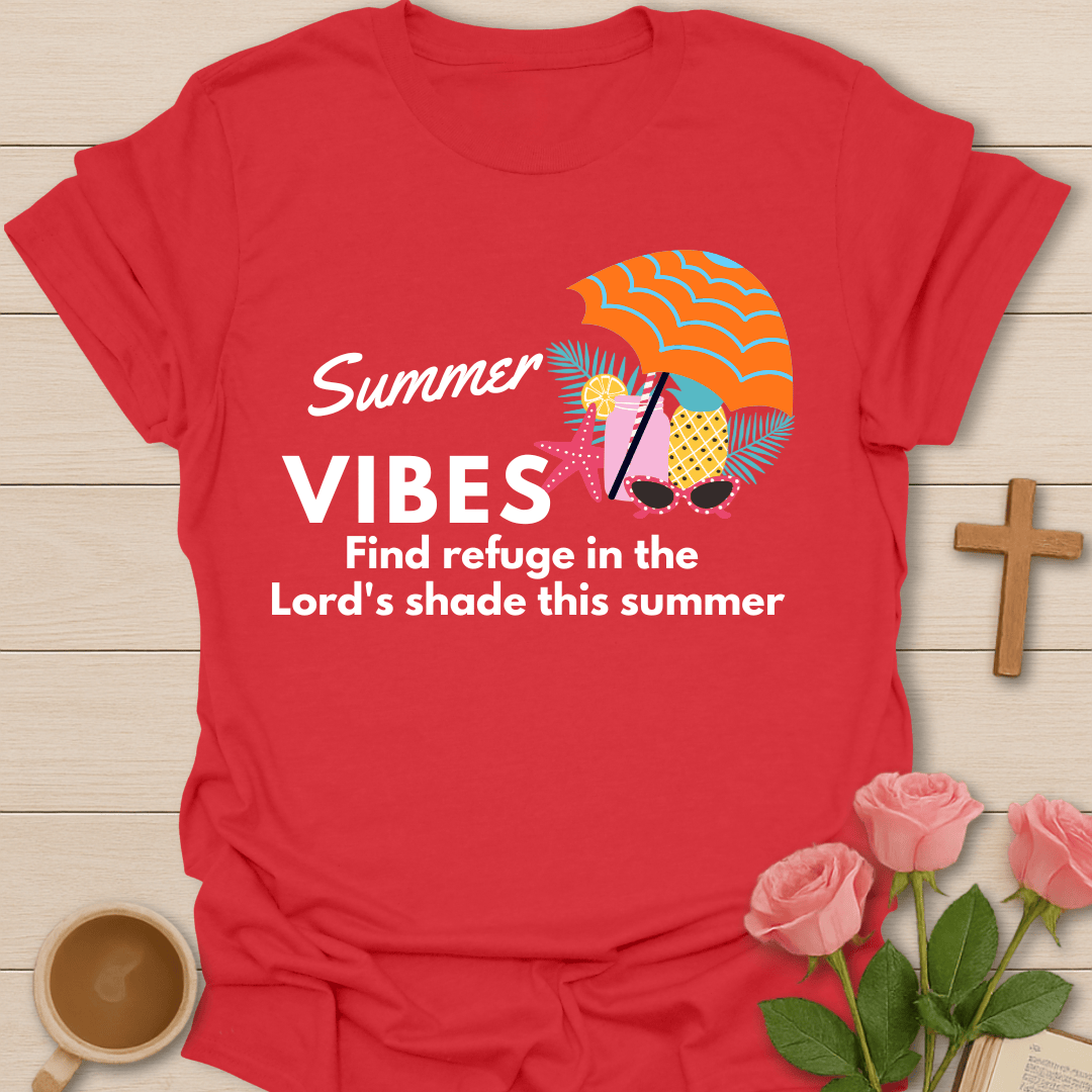 A red short-sleeve graphic T-shirt laid flat on a light wood surface, with a colorful summer-themed print reading 'Summer VIBES Find refuge in the Lord's shade this summer.' The design includes a striped orange and yellow umbrella, pink drink cups, sunglasses, and palm leaves.