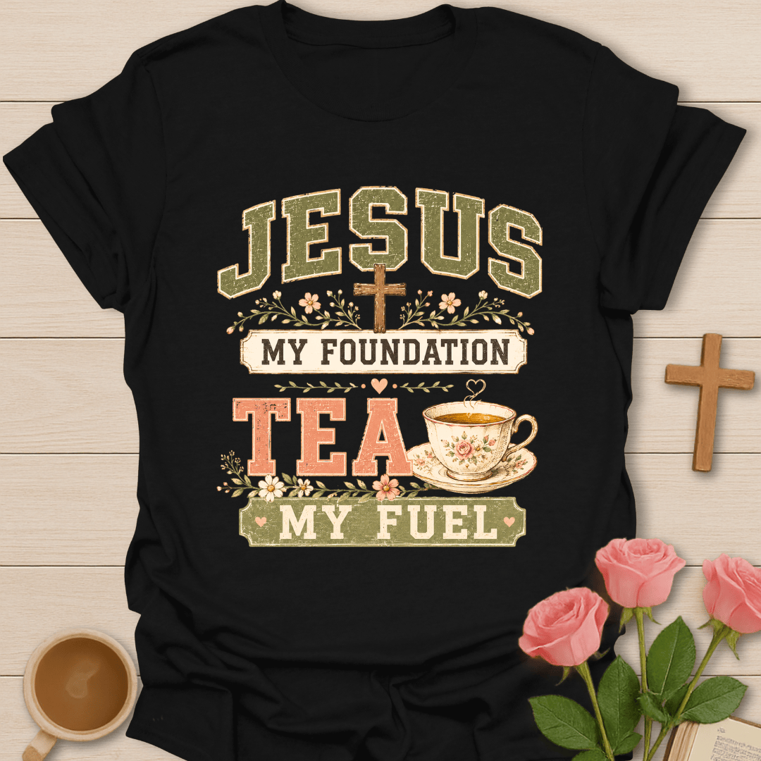 A black short-sleeve graphic T-shirt laid flat on a light wood surface, with decorative props including a small wooden cross, pink roses, and a teacup. The shirt graphic reads “JESUS MY FOUNDATION TEA MY FUEL” with floral and cross motifs.