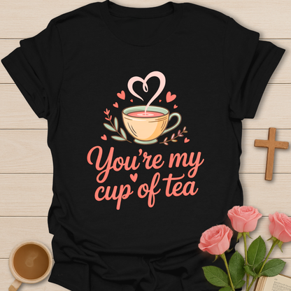 Black t-shirt with a graphic of a teacup and hearts, text 'You're my cup of tea', on a wooden surface with flowers and a coffee cup.