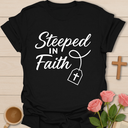 Black t-shirt with 'Steeped in Faith' text and cross graphic on a wooden surface with flowers and a cup.