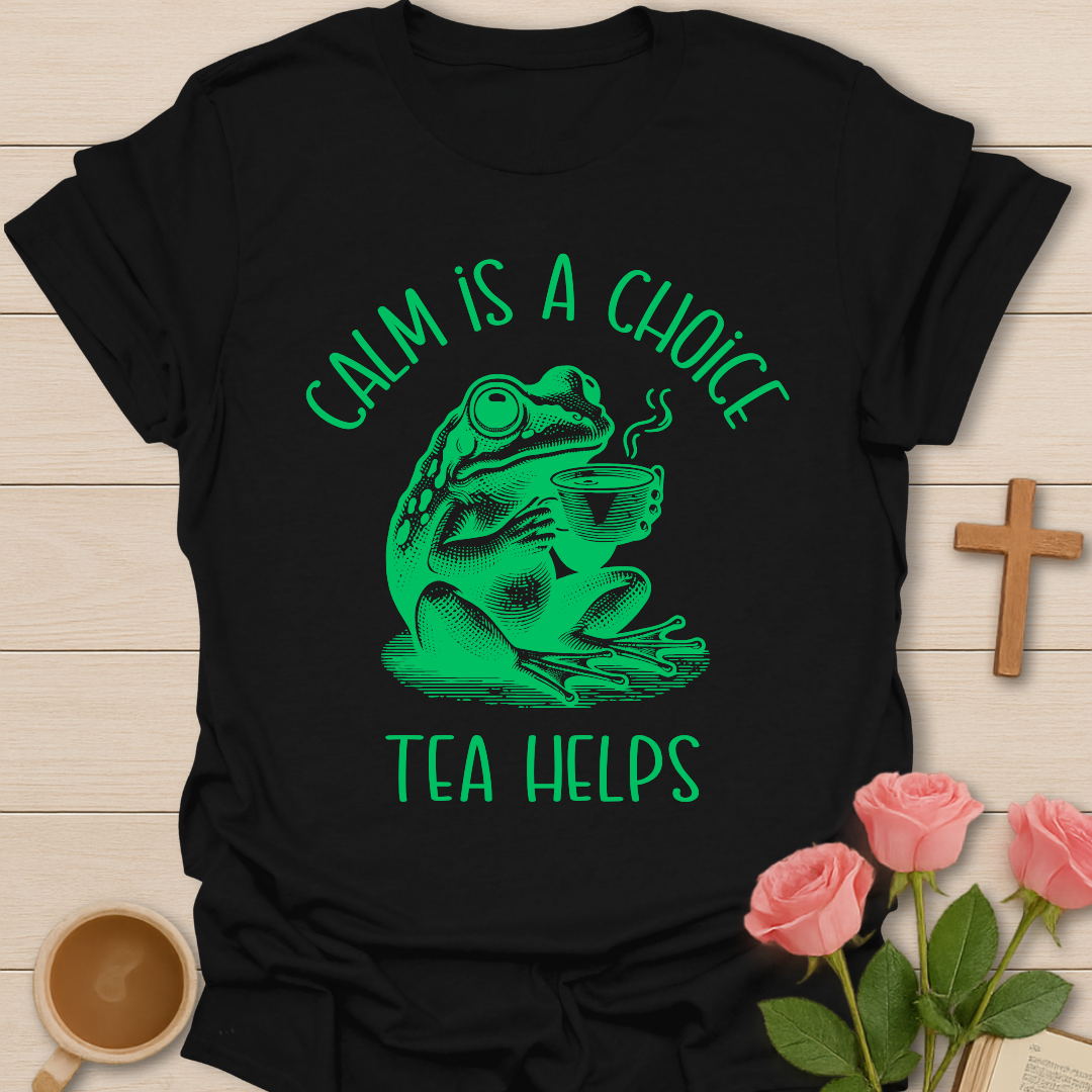 Black t-shirt with green frog graphic and text 'Calm is a choice, tea helps' on a wooden surface with flowers and a cup.
