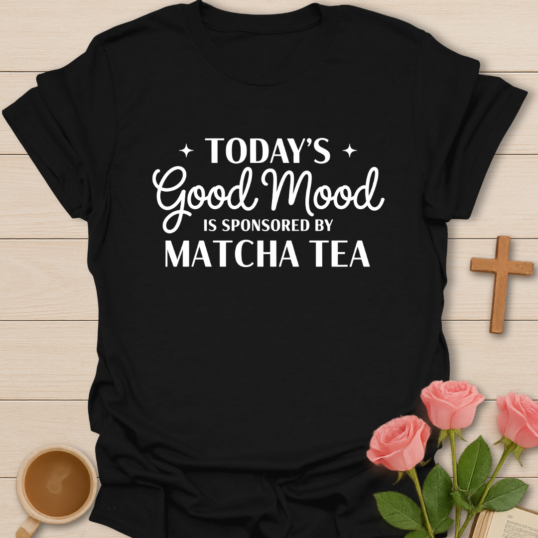 Black t-shirt with 'Today's Good Mood is Sponsored by Matcha Tea' text on a wooden surface with flowers and a cup.