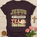 A dark brown graphic t-shirt laid flat on a light wood background, surrounded by decorative props including a wooden cross, pink roses, and a cup of tea. The shirt features a vintage-style graphic that reads 'JESUS MY FOUNDATION TEA MY FUEL' with a cross, floral details, and a teacup illustration.