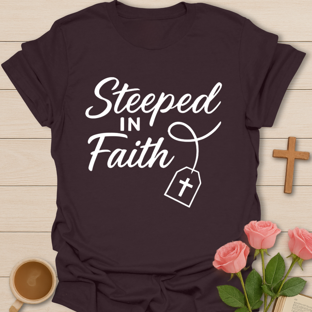 Brown t-shirt with 'Steeped in Faith' text and cross graphic on a wooden surface with flowers and a cup.