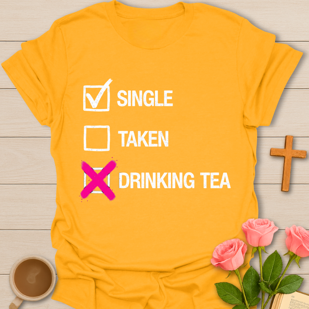 Yellow t-shirt with text 'SINGLE — TAKEN — DRINKING TEA' on a wooden surface with flowers and a cup.