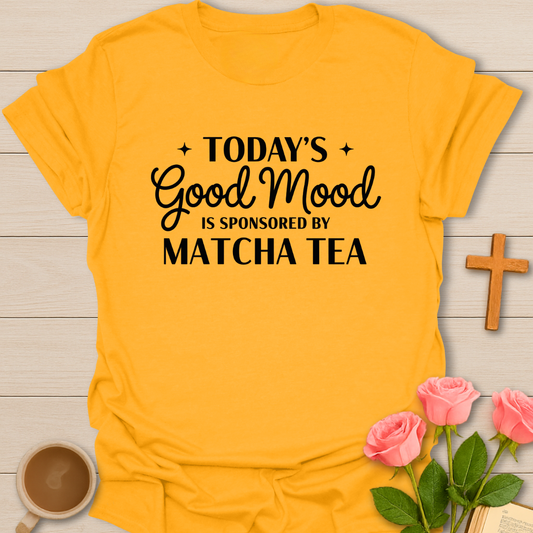 Yellow t-shirt with 'Today's Good Mood is Sponsored by Matcha Tea' text on a wooden surface with a cup of coffee and pink roses.