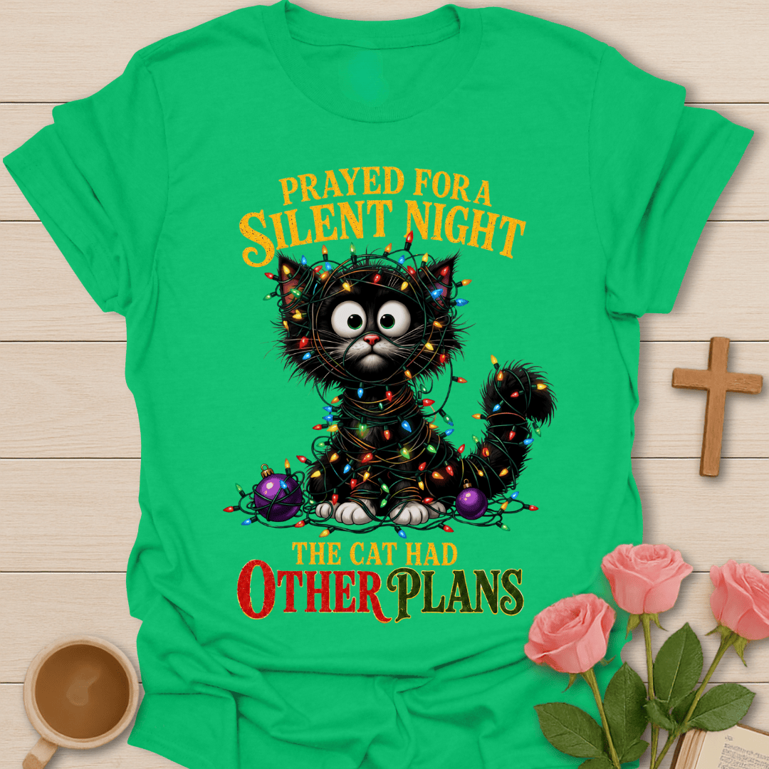 A bright green short-sleeve graphic T-shirt laid flat on a light wood surface, featuring a black cat surrounded by colorful Christmas lights and ornaments with the text 'PRAYED FOR A SILENT NIGHT THE CAT HAD OTHER PLANS.' Pink roses, a small wooden cross, and a cup are placed around the shirt as styling props.