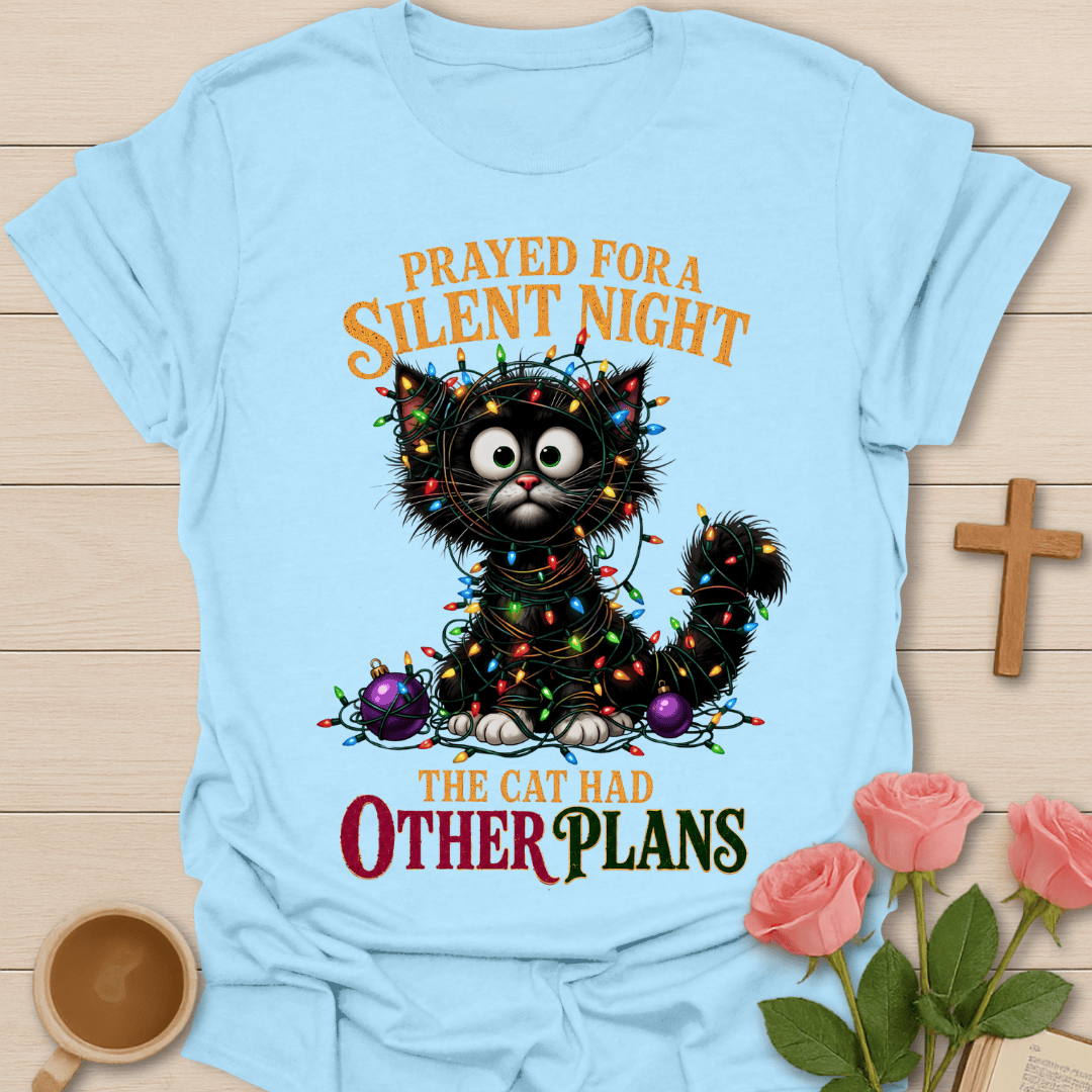 A light blue graphic t-shirt laid flat on a light wood background, featuring a black cat wrapped in Christmas lights with the text 'PRAYED FOR A SILENT NIGHT THE CAT HAD OTHER PLANS'. The shirt is styled with pink roses, a wooden cross, and a small cup.