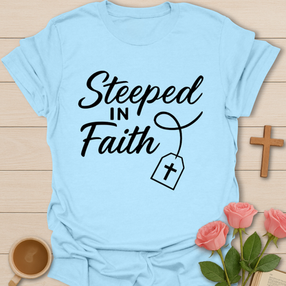 Light blue t-shirt with 'Steeped in Faith' text and a cross on a wooden surface with flowers and a cup.