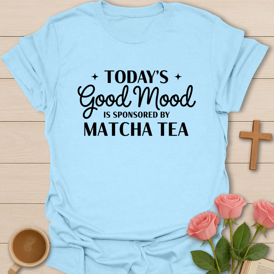 Light blue t-shirt with text 'Today's Good Mood is Sponsored by Matcha Tea' on a wooden surface with flowers and a cup.