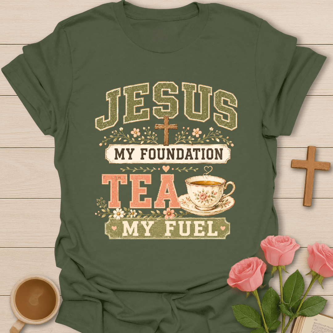 An olive green graphic t-shirt laid flat on a light wood background, surrounded by decorative props including pink roses, a wooden cross, and a cup of tea. The t-shirt features a vintage-style printed graphic that reads 'JESUS MY FOUNDATION TEA MY FUEL' with a cross, floral details, and a cup of tea.
