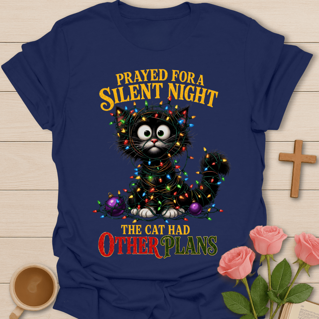 A navy blue graphic t-shirt laid flat on a light wood background, featuring a black cat wrapped in colorful Christmas lights with the text 'PRAYED FOR A SILENT NIGHT THE CAT HAD OTHER PLANS'. The shirt is styled with pink roses, a wooden cross, and a cup on a light wood surface.