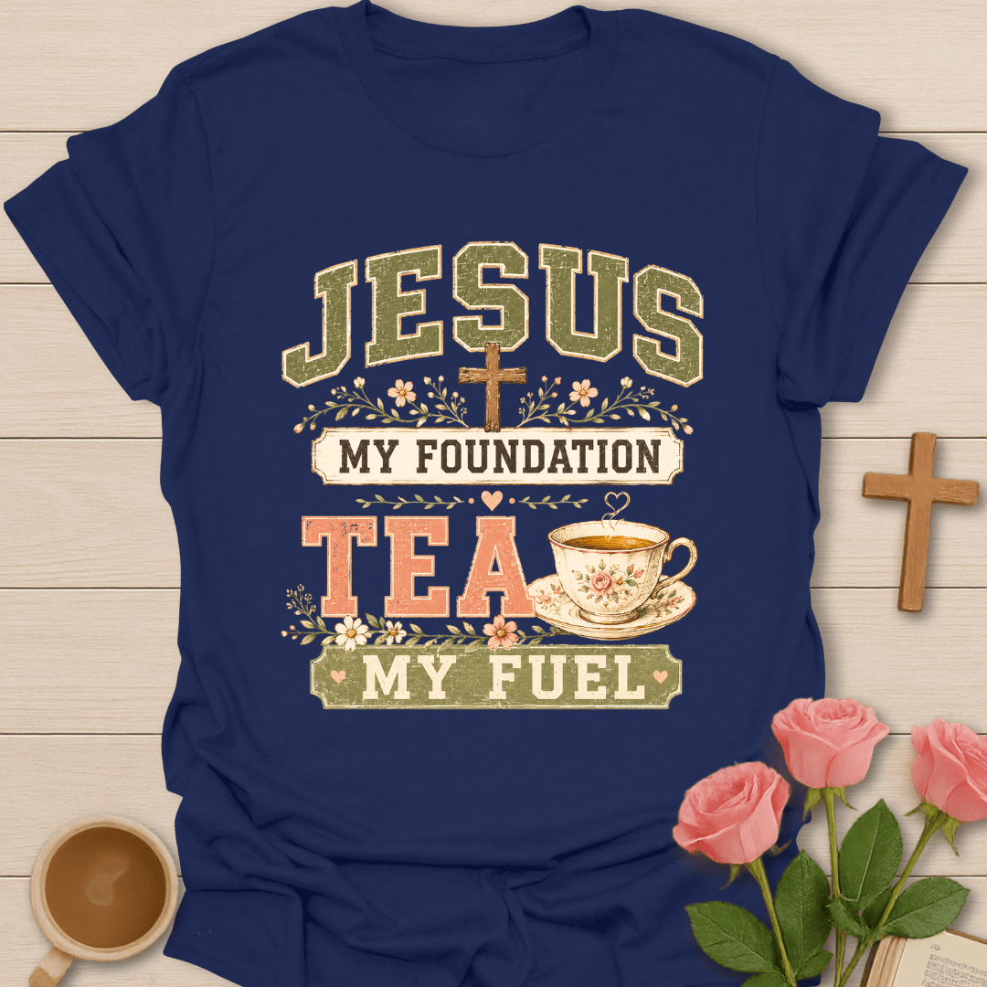 A navy blue graphic t-shirt laid flat on a light wood background, surrounded by decorative props including a wooden cross, pink roses, and a cup of tea. The t-shirt features a retro-style graphic that reads 'JESUS MY FOUNDATION TEA MY FUEL' with a small cross and floral details.