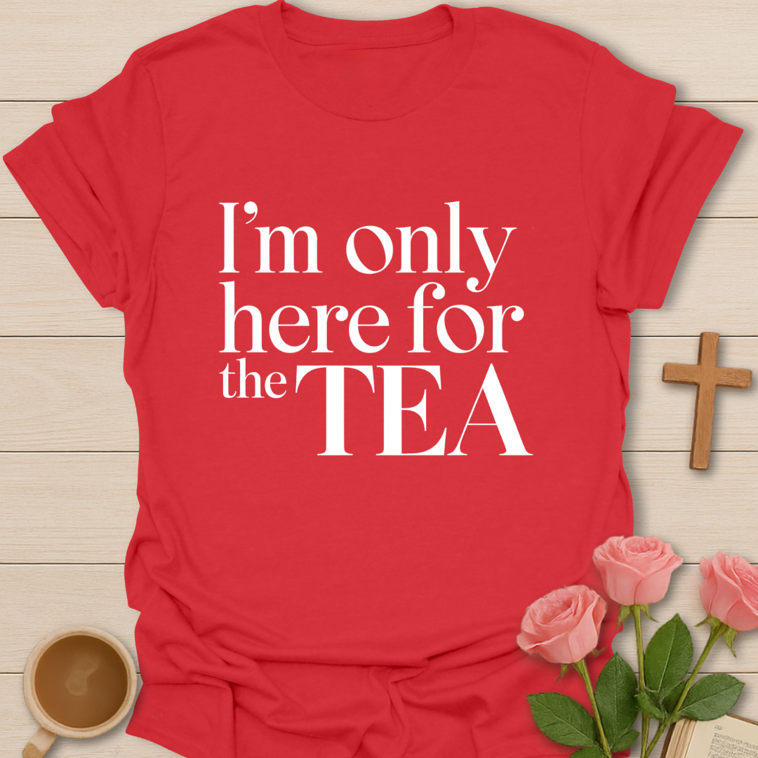 Red t-shirt with 'I'm only here for the TEA' text on a wooden surface with flowers and a cup.