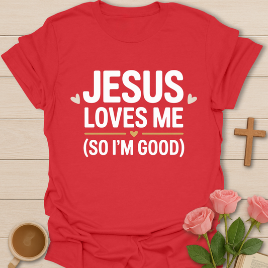 Red t-shirt with 'Jesus Loves Me (So I'm Good)' text on a wooden surface with flowers and a cross.