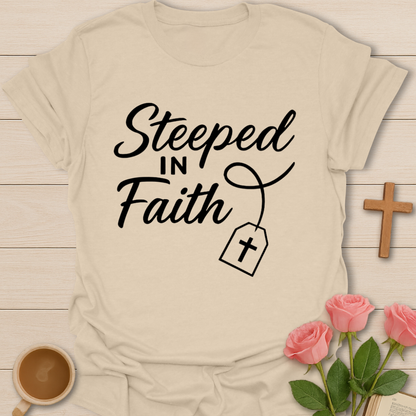 Beige t-shirt with 'Steeped in Faith' text on a wooden surface with flowers and a cross.