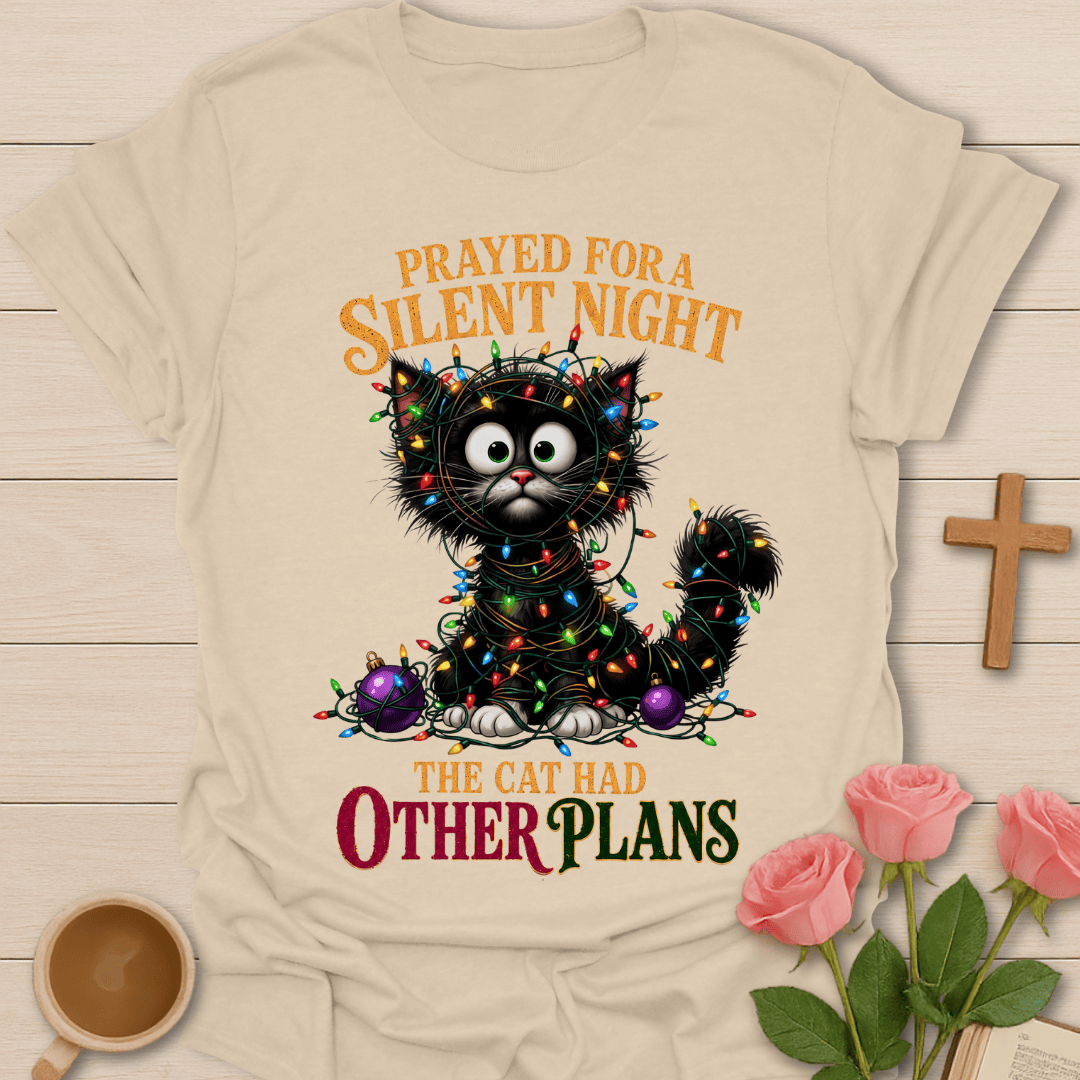 A beige graphic t-shirt laid flat on a light wood background, featuring a black cat wrapped in colorful Christmas lights with the text 'PRAYED FOR A SILENT NIGHT THE CAT HAD OTHER PLANS'. The scene is styled with pink roses, a wooden cross, and a cup of coffee.