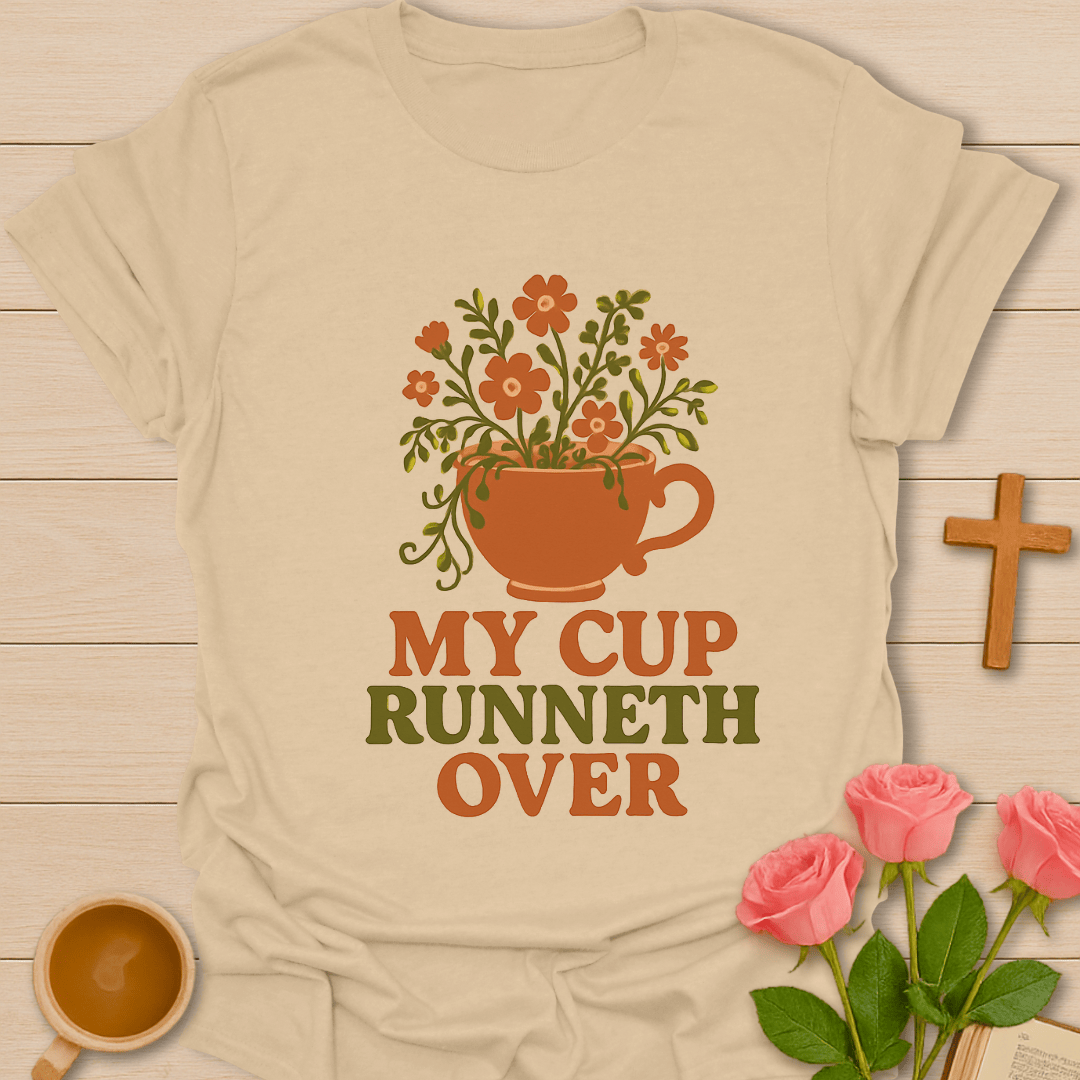 Beige t-shirt with floral cup design and 'My Cup Runneth Over' text on a wooden surface with flowers and a cross.