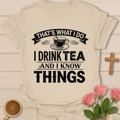 Beige t-shirt with black text 'That's what I do, I drink tea and I know things' on a wooden surface with a cup of tea and roses.