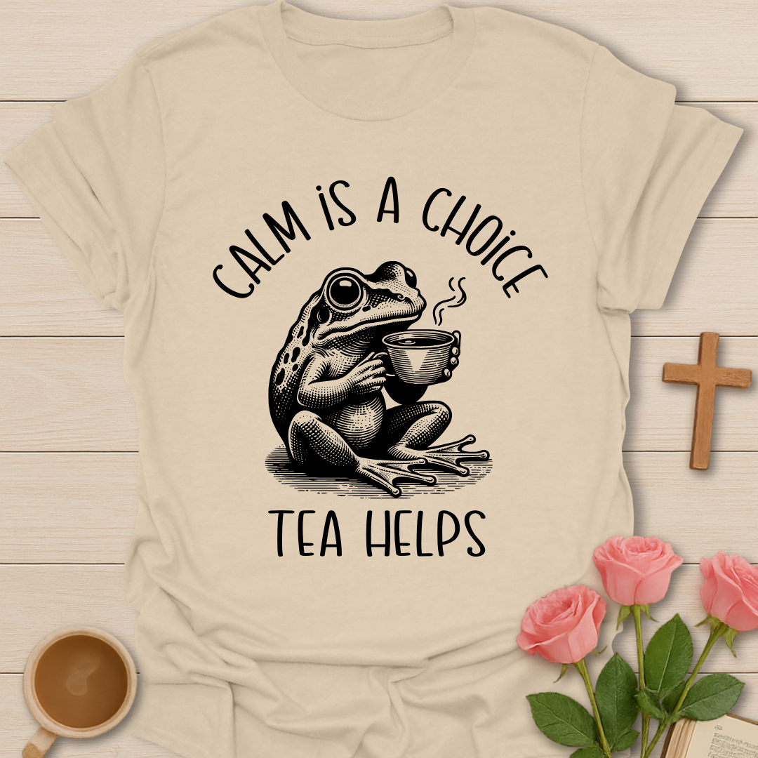 Beige t-shirt with a frog holding a tea cup and text 'Calm is a choice, tea helps' on a wooden surface with a cross and flowers.