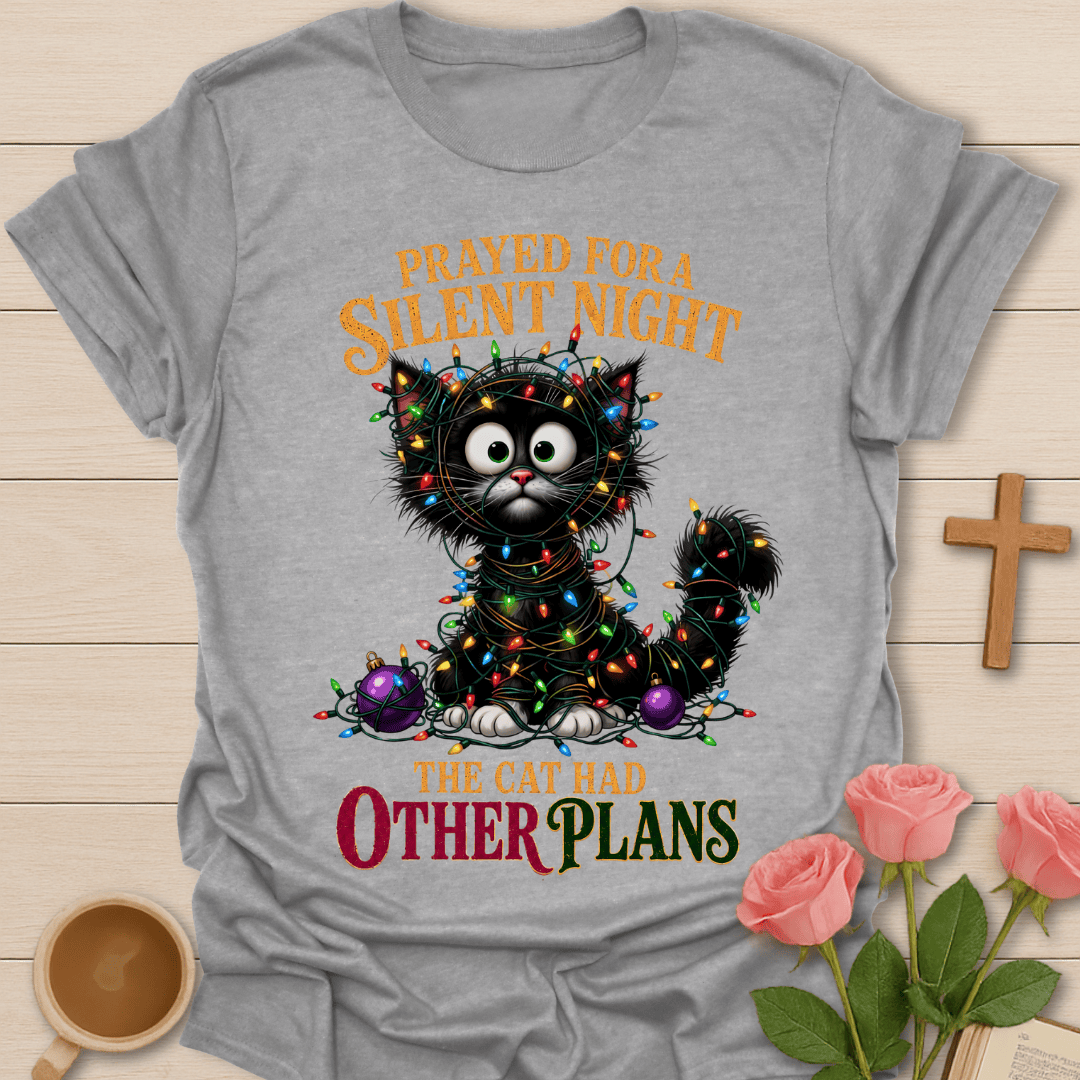 A light gray graphic t-shirt featuring a cartoon cat wrapped in Christmas lights, with the text 'PRAYED FOR A SILENT NIGHT THE CAT HAD OTHER PLANS'. The t-shirt is displayed on a light wood background with decorative props including a wooden cross, pink roses, and a coffee cup.