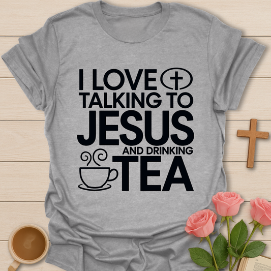 Gray t-shirt with text 'I love talking to Jesus and drinking tea' on a wooden surface with a cup, roses, and cross.