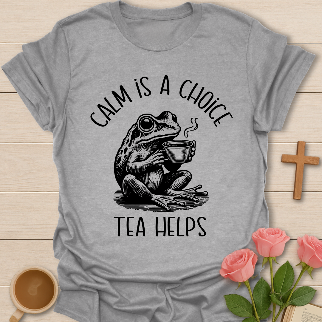 Gray t-shirt with a frog holding a tea cup graphic and text 'Calm is a choice, tea helps' on a wooden surface with flowers and a cross.
