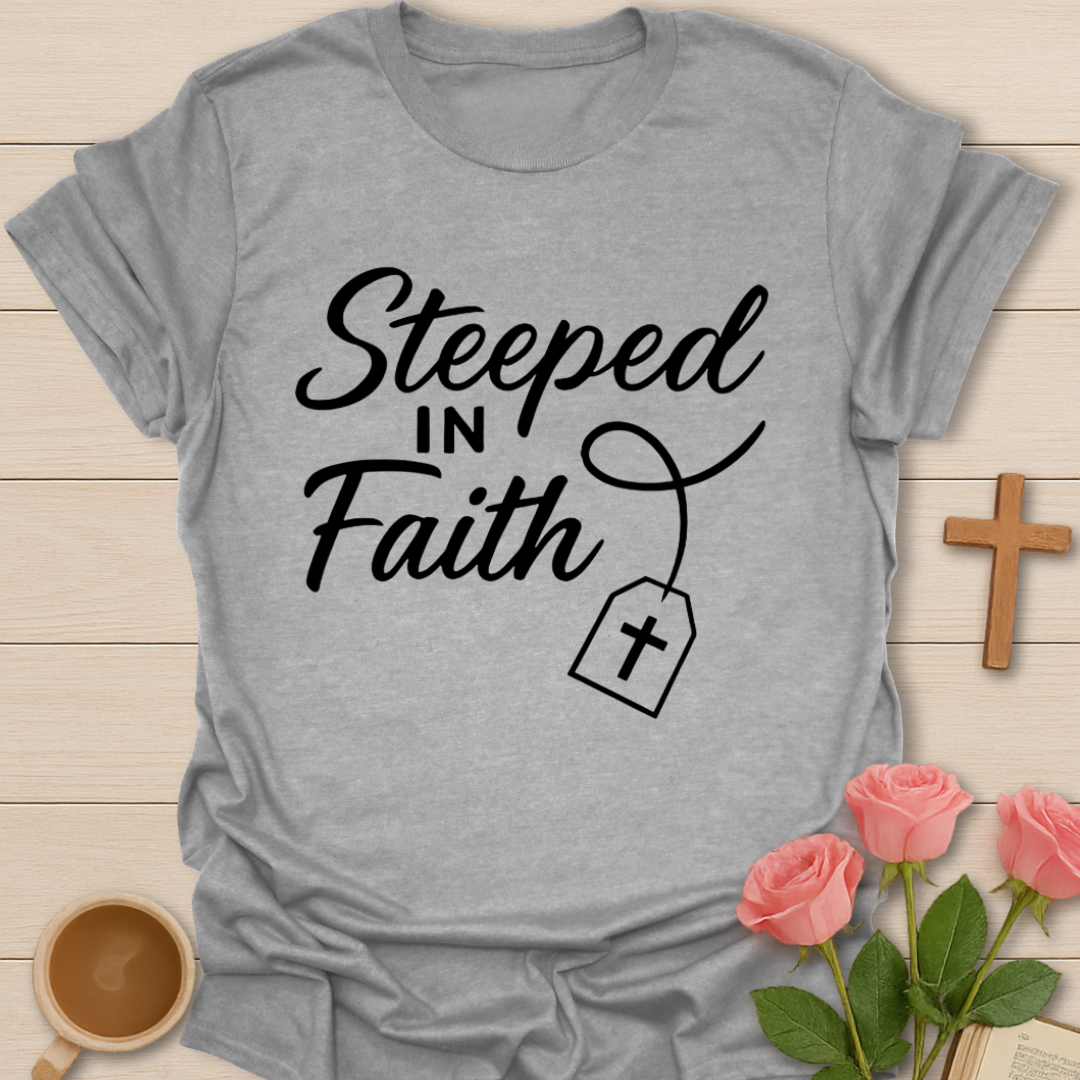 Gray t-shirt with 'Steeped in Faith' text on a wooden surface with flowers and a cross.