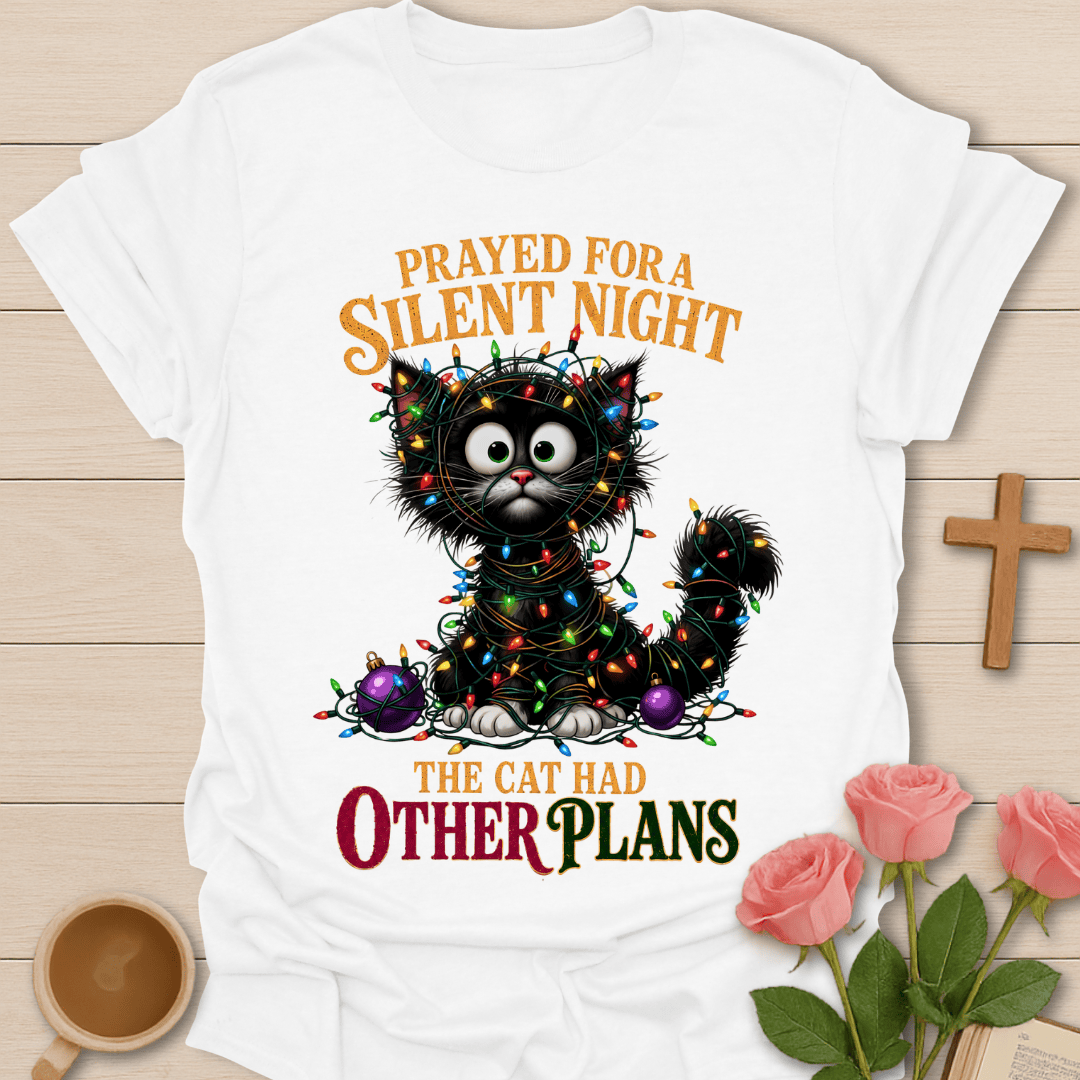 A white short-sleeve graphic T-shirt laid flat on a light wood surface, featuring a black cat surrounded by colorful Christmas lights and ornaments with the text 'PRAYED FOR A SILENT NIGHT THE CAT HAD OTHER PLANS.' Pink roses, a wooden cross, and a cup are placed around the shirt as styling props.