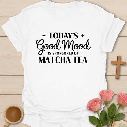 White t-shirt with 'Today's Good Mood is Sponsored by Matcha Tea' text on a wooden surface with flowers and a cup.