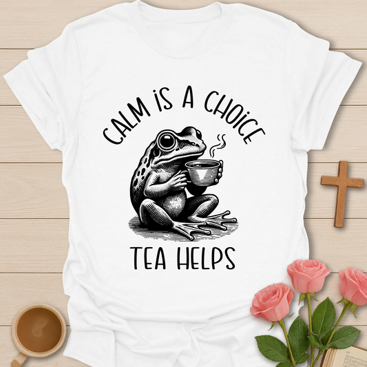 White t-shirt with a frog holding a tea cup graphic and text 'Calm is a choice, tea helps' on a wooden surface with flowers and a cross.