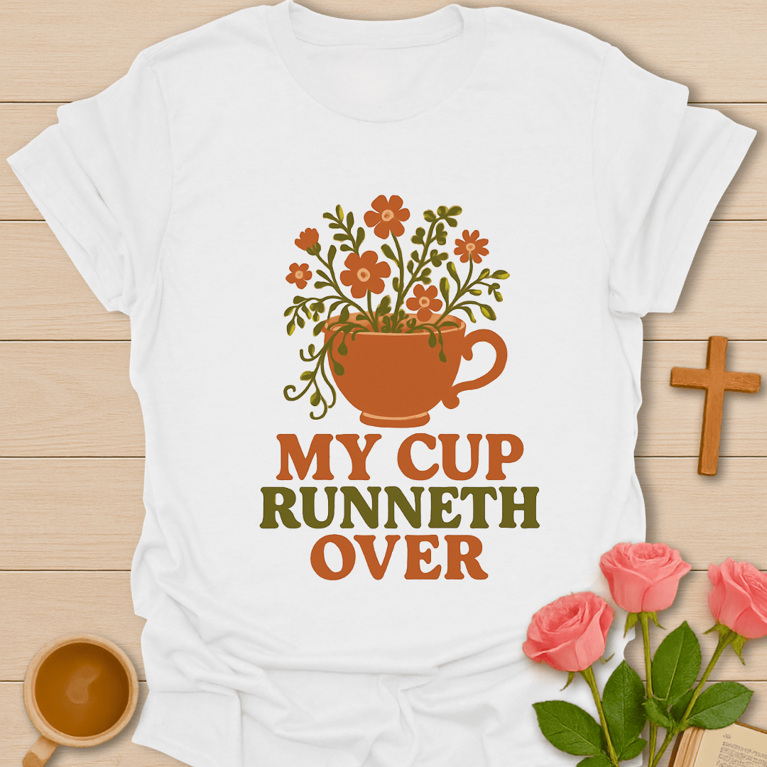 White t-shirt with floral cup design and 'My Cup Runneth Over' text on a wooden surface with flowers and a cross.