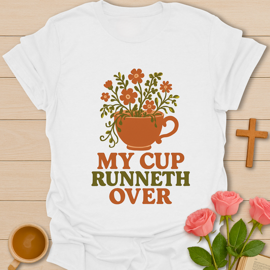 White t-shirt with floral cup design and 'My Cup Runneth Over' text on a wooden surface with flowers and a cross.