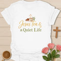 Jesus Tea and a Quiet Life t-shirt with gold and sage script, a closed Bible and a teacup with cross-shaped steam. - White