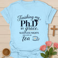 PhD By Grace shirt with large navy PhD lettering, graduation cap, script grace text, sleepless nights phrase, and teacup illustration. - Light Blue