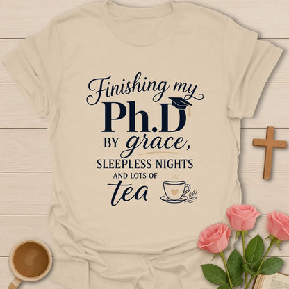 PhD By Grace shirt with large navy PhD lettering, graduation cap, script grace text, sleepless nights phrase, and teacup illustration. - Sand