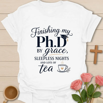 PhD By Grace shirt with large navy PhD lettering, graduation cap, script grace text, sleepless nights phrase, and teacup illustration. - White