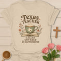 Texas Teacher shirt with sage teacup on open Bible, Texas silhouette, wildflowers, distressed brown lettering, and vintage boutique colors. - Sand
